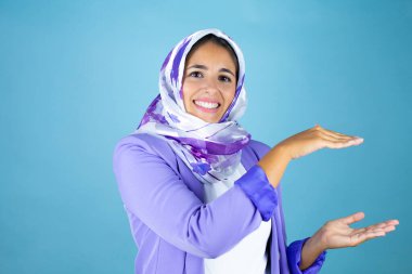 Genç, güzel bir Arap kadın, izole edilmiş mavi arka plan üzerine tesettür takıyor. Ellerinde büyük ve büyük bir işaret, ölçü sembolü var. Kameraya bakıp gülümsüyor. Ölçüm konsepti.