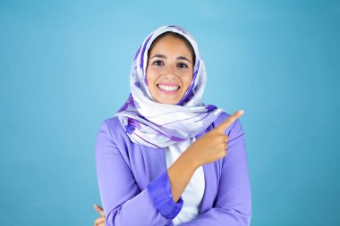 Genç, güzel bir Arap kadın, izole edilmiş mavi arka plan üzerine tesettür takmış, gülümsüyor ve elini ve parmağını kenarı işaret ediyor.