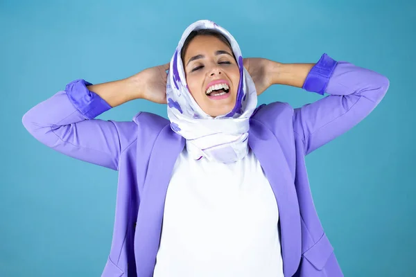 若いです美しいですArab女性身に着けていますイスラムヒジャーブ上の隔離された青の背景リラックスしてストレッチ 腕と手後ろ頭と首笑顔幸せな — ストック写真