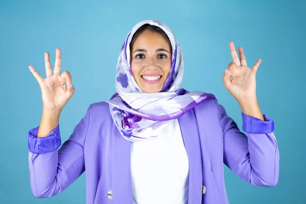 若いです美しいですアラブ女性身に着けていますイスラムヒジャーブ上の隔離された青の背景を行いますOkサインと指と笑顔 優れたシンボル — ストック写真
