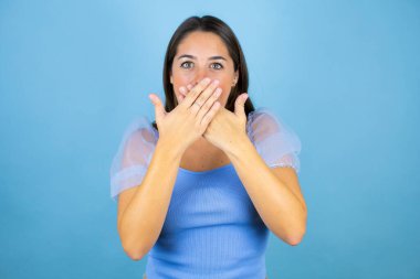 Soyutlanmış mavi arka planda genç ve güzel bir kadın yanlışlıkla ağızlarını kapatarak şok geçirdi. Gizli konsept.