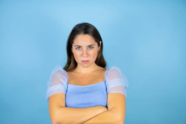 Genç, güzel bir kadın, izole edilmiş mavi arka planda şüpheci ve gergin, yüzünde çapraz kollu onaylanmamış bir ifade var.