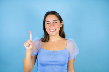 İzole edilmiş mavi arka planda genç ve güzel bir kadın bir numaralı parmaklarıyla işaret ederken kendinden emin ve mutlu bir şekilde gülümsüyor.