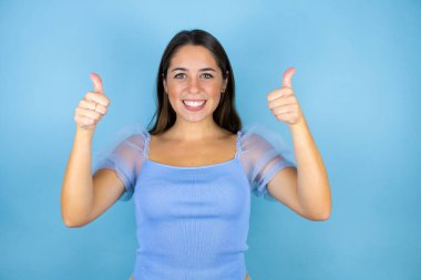 İzole edilmiş mavi arka planda genç ve güzel bir kadın gülümsüyor ve baş parmaklarıyla işaret veriyor.