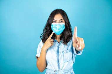 Tecrit edilmiş mavi arka planda tıbbi maske takan genç ve güzel bir kadın. Yüzünde olumsuz ve ciddi bir jest olan uyarı ifadesi.