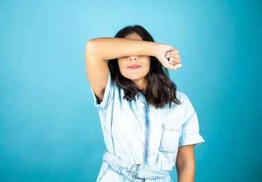 İzole edilmiş mavi arka planda kot tulum giyen güzel bir kadın gözlerini tek koluyla kapatıyor..