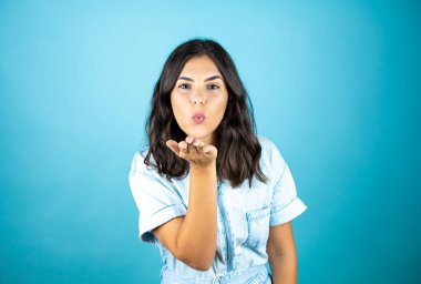 İzole edilmiş mavi arka plan üzerinde kot tulum giyen genç ve güzel bir kadın havayla öpüşürken kameraya bakıyor. Güzel ve seksi. Aşk ifadesi.