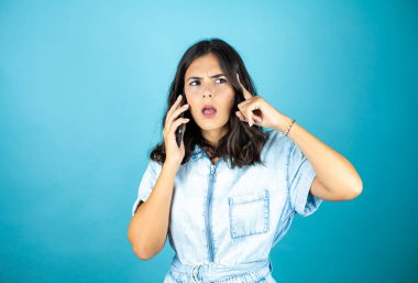 İzole edilmiş mavi arka planda kot pantolon giyen güzel bir kadın telefonda kafası karışık bir ifadeyle konuşuyor..