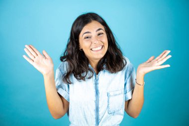 İzole edilmiş mavi arka plan üzerinde kot tulum giyen genç ve güzel bir kadın. Kolları ve elleri havada, kafası karışmış bir ifade..