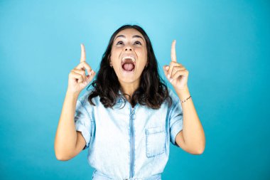 İzole edilmiş mavi arka planda kot pantolon giyen genç ve güzel bir kadın şaşkın şaşkın bakıyor ve parmaklarıyla işaret ediyor ve kollarını kaldırıyordu..