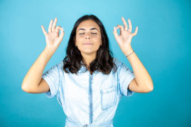 İzole edilmiş mavi arka planda kot pantolon giyen genç ve güzel bir kadın rahatla ve gözleri kapalı gülümse. Parmaklarıyla meditasyon hareketi yapıyor..