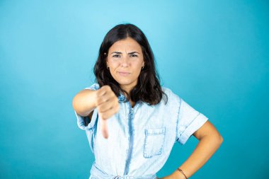 İzole edilmiş mavi arka planda kot pantolon giyen genç ve güzel bir kadın. Kızgın bir yüzü var..