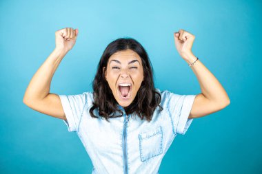 İzole edilmiş mavi arka planda kot pantolon giyen genç ve güzel bir kadın mutlu ve heyecanlı. Kollarını kaldırıp, gülümseyerek ve başarı için çığlık atarak kazanan bir jest yapıyor..