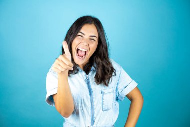 İzole edilmiş mavi arka planda kot pantolon giyen güzel bir kadın gülümsüyor ve baş parmağıyla işaret veriyor..