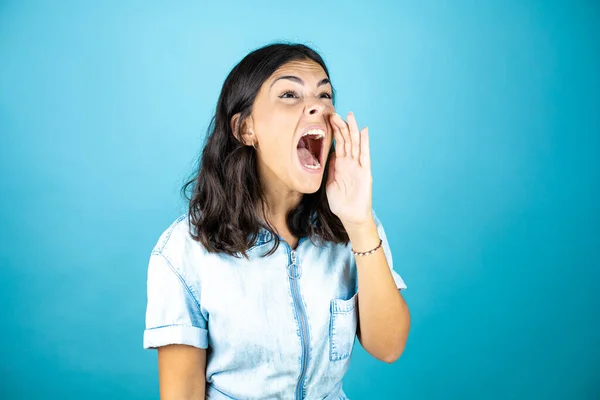 Young beautiful woman wearing a denim jumpsuit over isolated blue background shouting and screaming loud to side with hands on mouth.