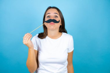 Soyutlanmış mavi arka planda genç ve güzel bir kadın bıyık maskesiyle mutlu.