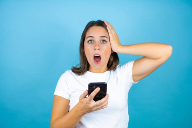 Soyutlanmış mavi arka planda sohbet eden ve telefonuyla şaşıran genç ve güzel bir kadın.