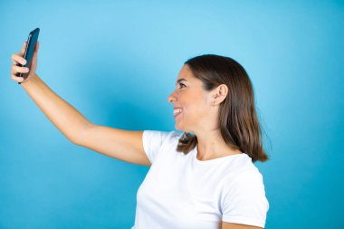 İzole edilmiş mavi arka planda genç ve güzel bir kadın telefonuyla selfie çekiyor.