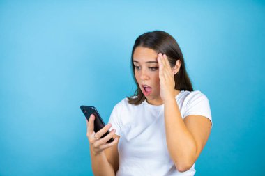 Soyutlanmış mavi arka planda genç ve güzel bir kadın. Konuşuyor, şaşırıyor ve telefonuyla bakıyor.