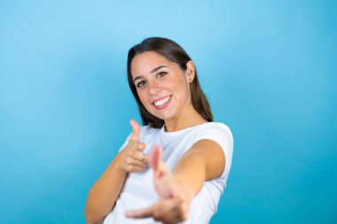 Soyutlanmış mavi arka planda genç ve güzel bir kadın seni ve kamerayı işaret ediyor, parmaklarıyla, mutlu ve mutlu gülümsüyor.
