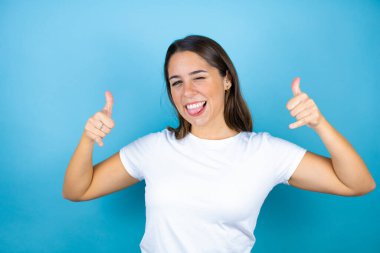 İzole edilmiş mavi arka planda genç ve güzel bir kadın çılgın bir ifadeyle el kaldırarak rock sembolü yapıyor.