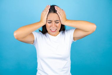 Genç ve güzel bir kadın, mavi arka planda, baş ağrısı, çaresiz ve stresli, çünkü elleri başının üstünde acı ve migren var.