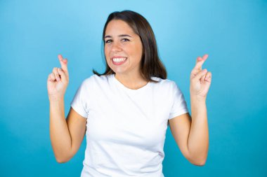 İzole edilmiş mavi arka plan üzerinde genç ve güzel bir kadın. Umut ve bakışıyla parmaklarını çapraz yapıyor.