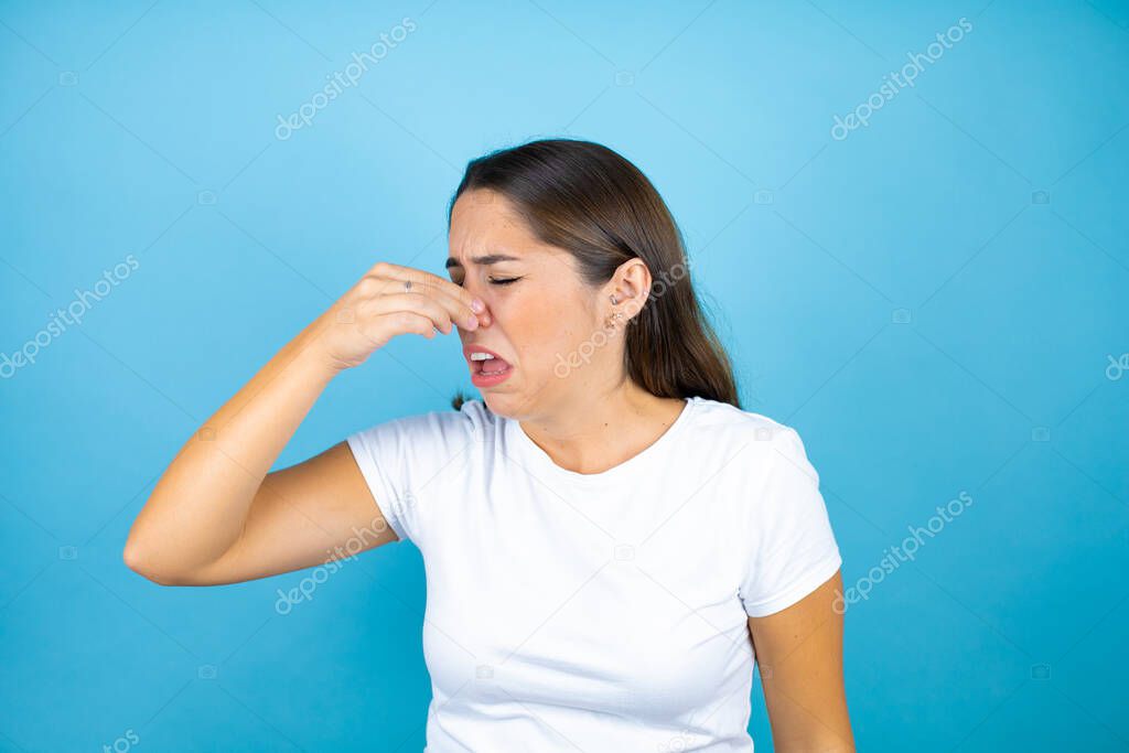 Young beautiful woman over isolated blue background smelling something stinky and disgusting, intolerable smell, holding breath with fingers on nose