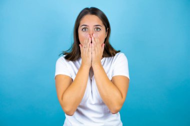 Genç ve güzel bir kadın, soyutlanmış mavi arka planda, elleri ağzında ve şaşırmış bir şekilde.