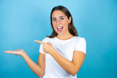 Genç ve güzel bir kadın, izole edilmiş mavi arka planda şaşırmış, elindeki bir şeyi gösteriyor ve işaret ediyor.