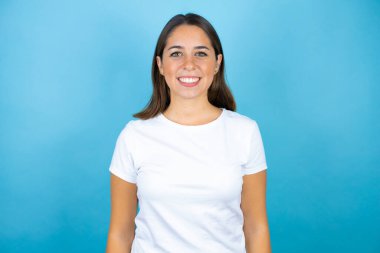 İzole edilmiş mavi arka planda genç ve güzel bir kadın gülümseyip kameraya bakıyor.