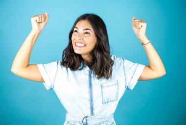 İzole edilmiş mavi arka planda kot pantolon giyen genç ve güzel bir kadın mutlu ve heyecanlı. Kollarını kaldırıp, gülümseyerek ve başarı için çığlık atarak kazanan bir jest yapıyor..