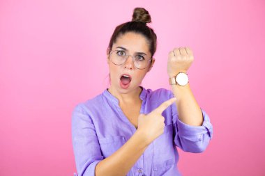 İzole edilmiş pembe arka planda gözlük takan genç ve güzel bir kadın şaşırdı ve saatini işaret etti çünkü geç oldu.