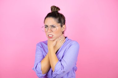 İzole edilmiş pembe arka planda gözlük takan genç ve güzel bir kadın ağrılı boyuna dokunuyor, boğaz ağrısı, grip, mantar ve enfeksiyon için.