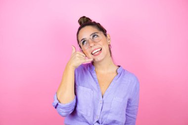 İzole edilmiş pembe arka planda gözlük takan güzel bir kadın gülümsüyor telefonla el ve parmaklarıyla jest yapıyor telefonda konuşuyor gibi.