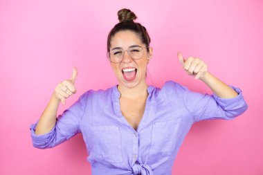 İzole edilmiş pembe arka planda gözlük takan genç ve güzel bir kadın çılgın bir ifadeyle el kaldırarak rock sembolü yapıyor.