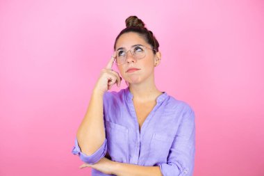 İzole edilmiş pembe arka planda gözlük takan genç ve güzel bir kadın. Gülümsüyor ve parmağıyla bir fikri olduğunu düşünüyor..