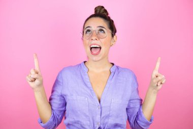 İzole edilmiş pembe arka planda gözlük takan genç ve güzel bir kadın şaşırmış ve şaşırmış bir şekilde yukarı bakıyor ve parmaklarıyla işaret ediyor ve kollarını kaldırıyor.