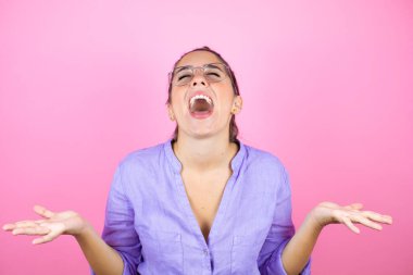 İzole edilmiş pembe arka planda gözlük takan genç ve güzel bir kadın. Kolları ve elleri havada, kafası karışmış bir ifade.