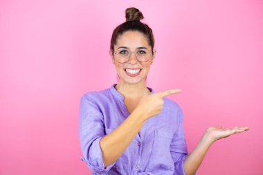 İzole edilmiş pembe arka planda gözlük takan genç ve güzel bir kadın elindeki bir şeyi gösterip işaret ediyor.