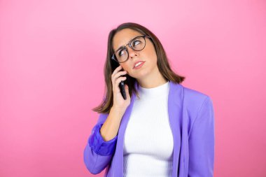 Genç ve güzel bir iş kadını pembe arka planda telefonda kafası karışık bir ifadeyle konuşuyor.