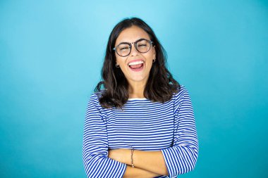 Kollarını kavuşturmuş gülümseyen mavi arka planda duran gözlüklü güzel kadın.