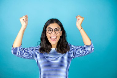 Gözlüklü güzel bir kadın mavi arka planda mutlu ve heyecanlı kollarını kaldırmış, gülümseyen ve başarı için bağıran, kazanan bir jest yapıyor..