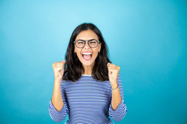 Gözlüklü güzel bir kadın mavi arka planda mutlu ve heyecanlı kollarını kaldırmış, gülümseyen ve başarı için bağıran, kazanan bir jest yapıyor..