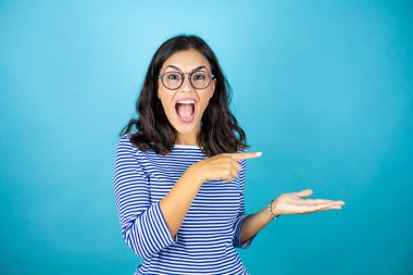 Gözlük takan güzel bir kadın mavi arka planda duruyor. Elindeki boşluğu hayret dolu bir ifadeyle gösteriyor..