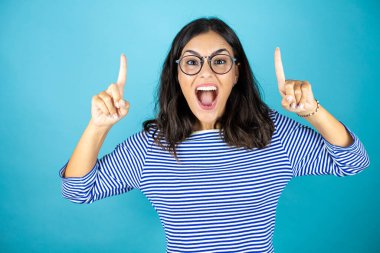 Gözlüklü güzel bir kadın mavi arka planda dikiliyor kameraya bakıyor ve parmaklarıyla işaret ediyor ve kollarını kaldırıyor.
