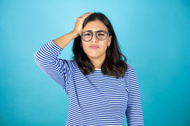 Mavi arka planda duran gözlüklü güzel bir kadın bir elini kafasına koyup sanki bir şey unutmuş gibi gülümsüyor..