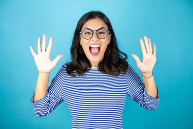Gözlüklü güzel bir kadın mavi arka planda duruyor ve parmaklarını on numarayla gösteriyor ve gülümsüyor kendinden emin ve mutlu