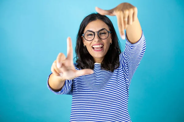 Gözlüklü güzel bir kadın mavi arka planda duruyor gülümsüyor elleri ve parmakları gülen yüzlü bir çerçeve yapıyor..