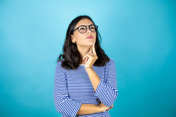 Gözlüklü güzel kadın mavi arka planda dikilip düşünceli bir şekilde yan tarafa bakıyor..